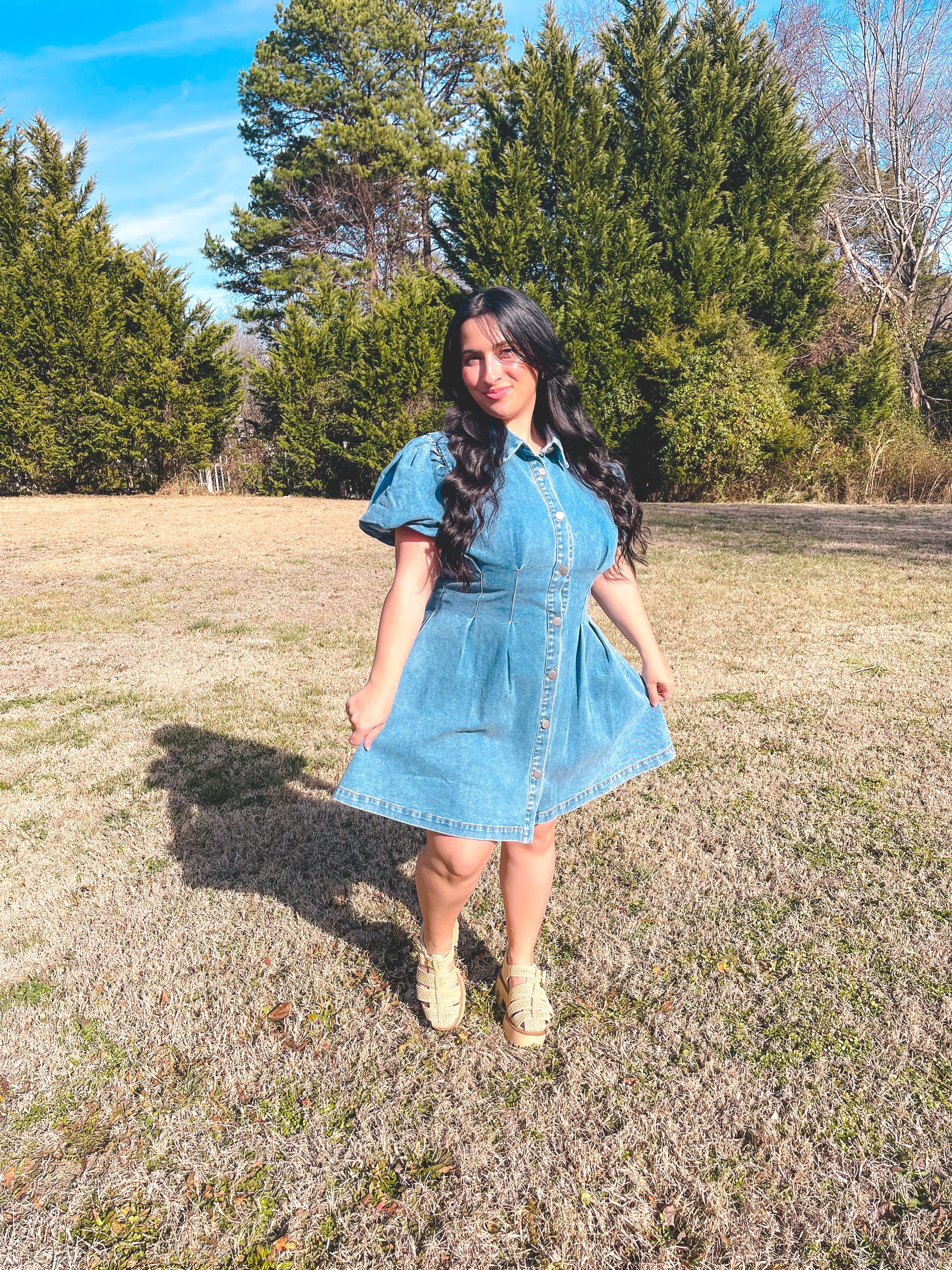 Woman in a denim dress standing in a field with trees in the background
