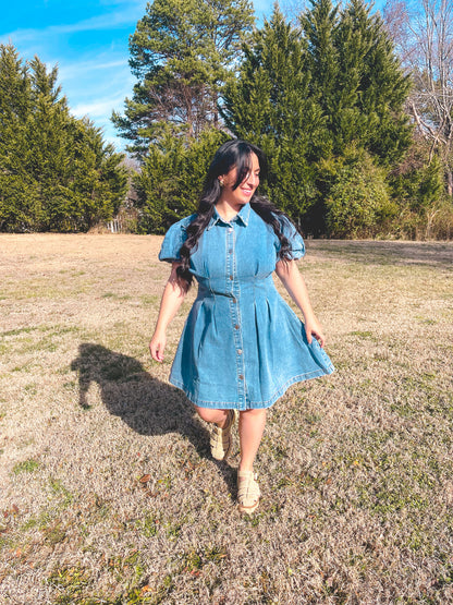 Woman in a denim dress standing in a field with trees in the background