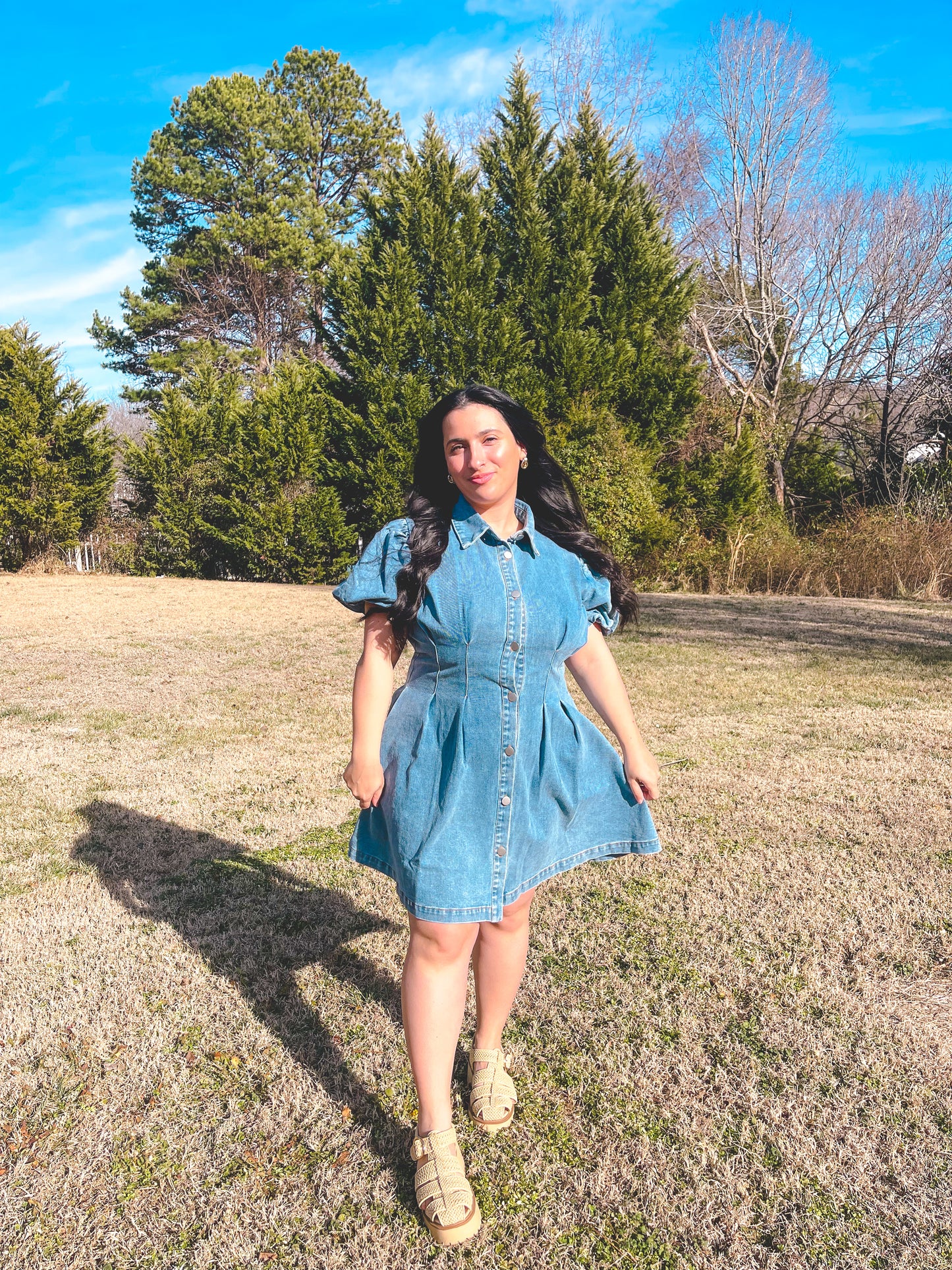 Woman in a denim dress standing in a field with trees in the background