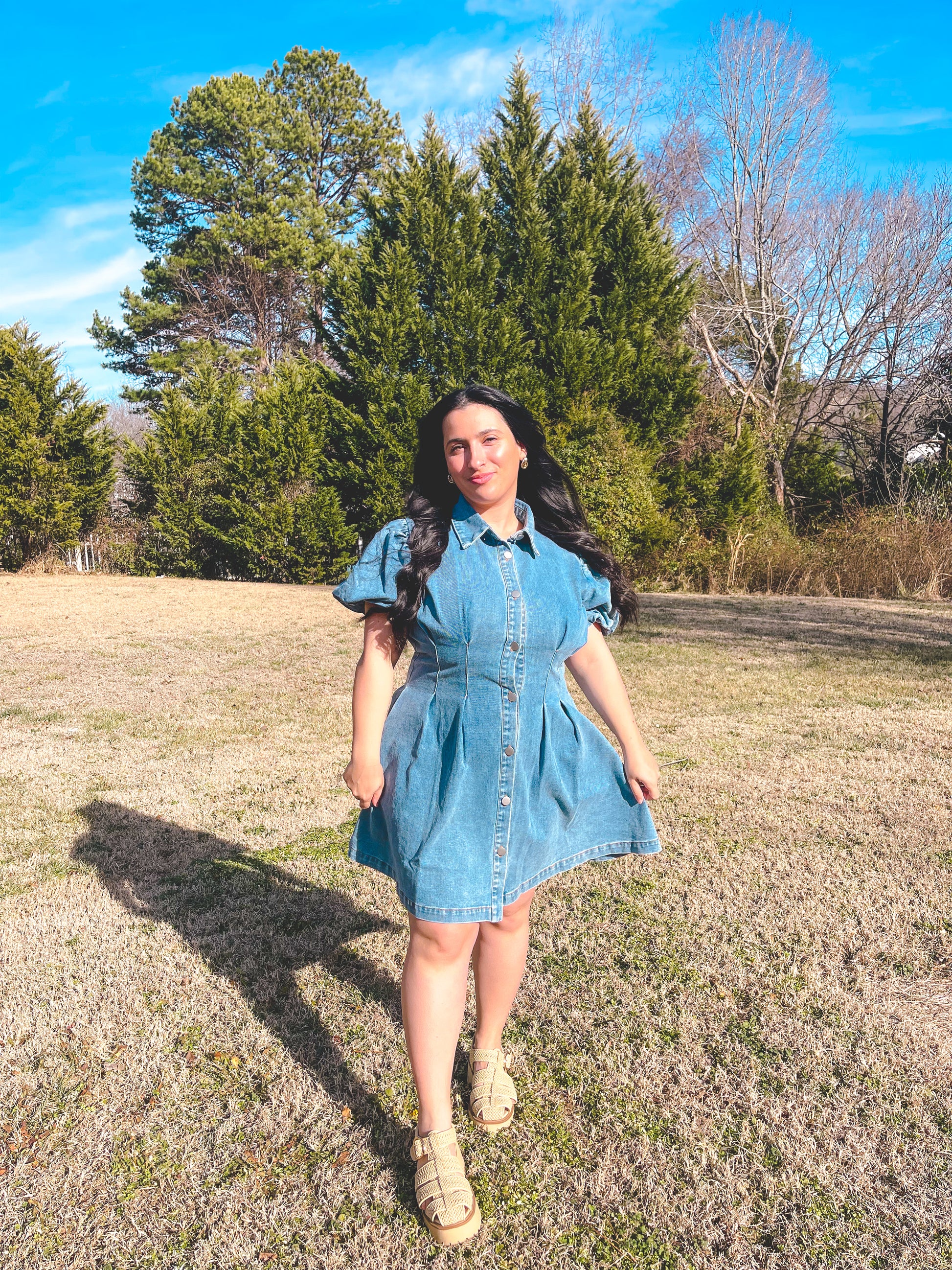 Woman in a denim dress standing in a field with trees in the background