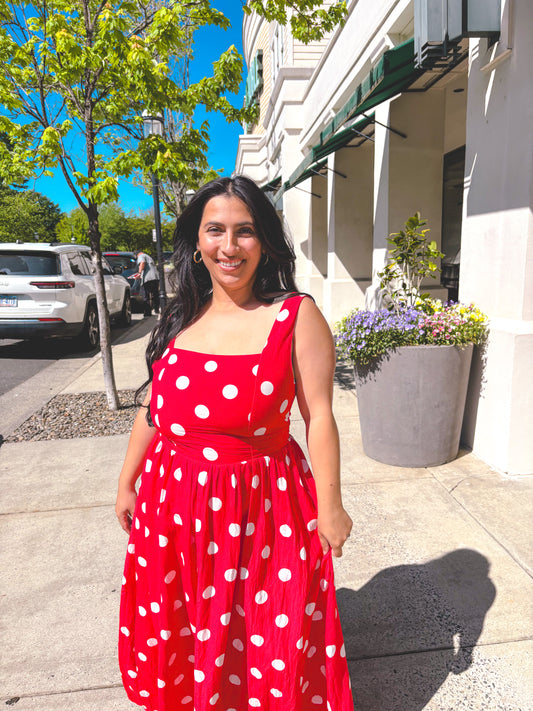 Woman in a red polka dot dress standing on a sidewalk with buildings and trees in the background.