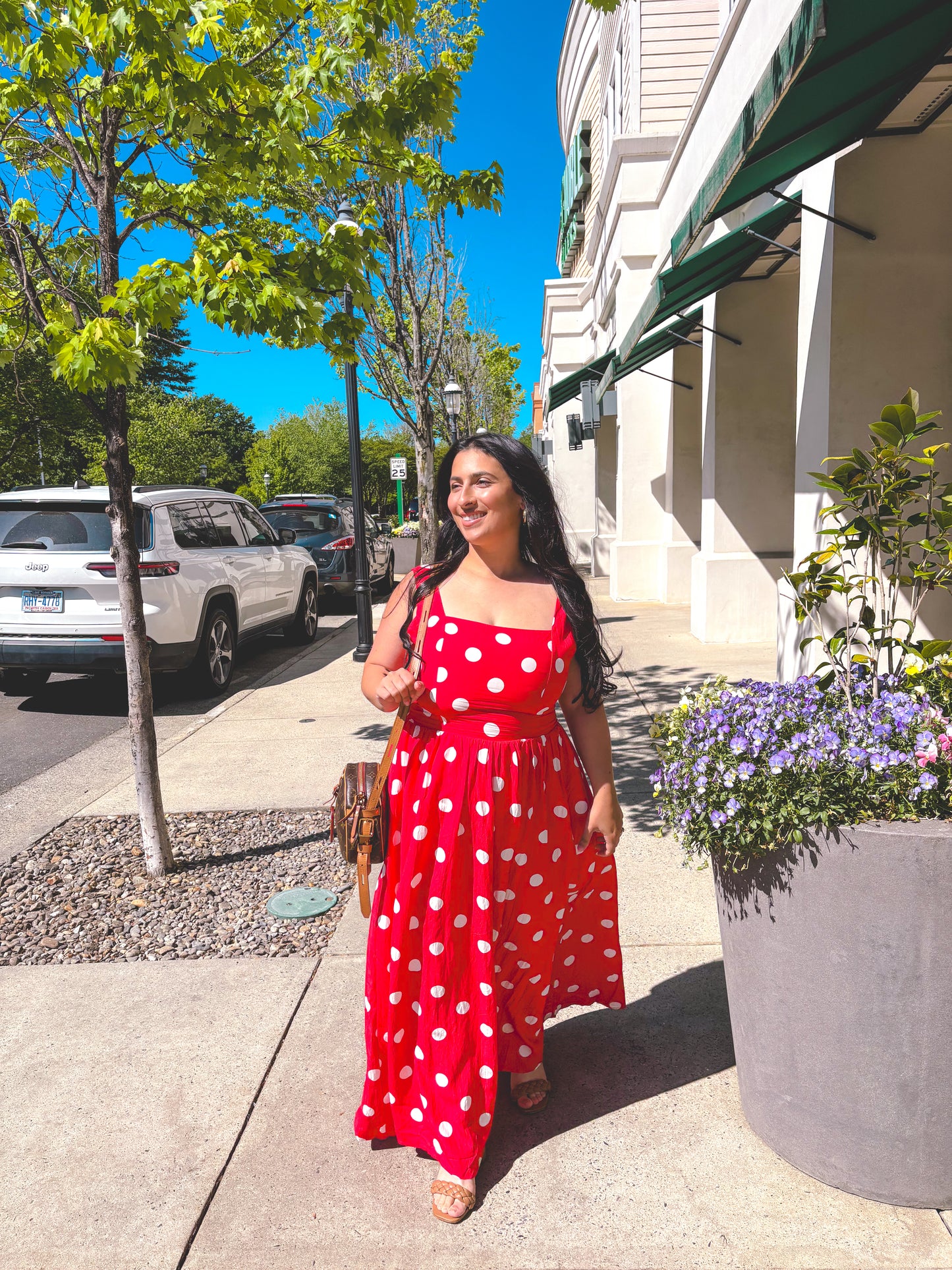 Woman in a red polka dot dress standing on a sidewalk with trees and buildings in the background.