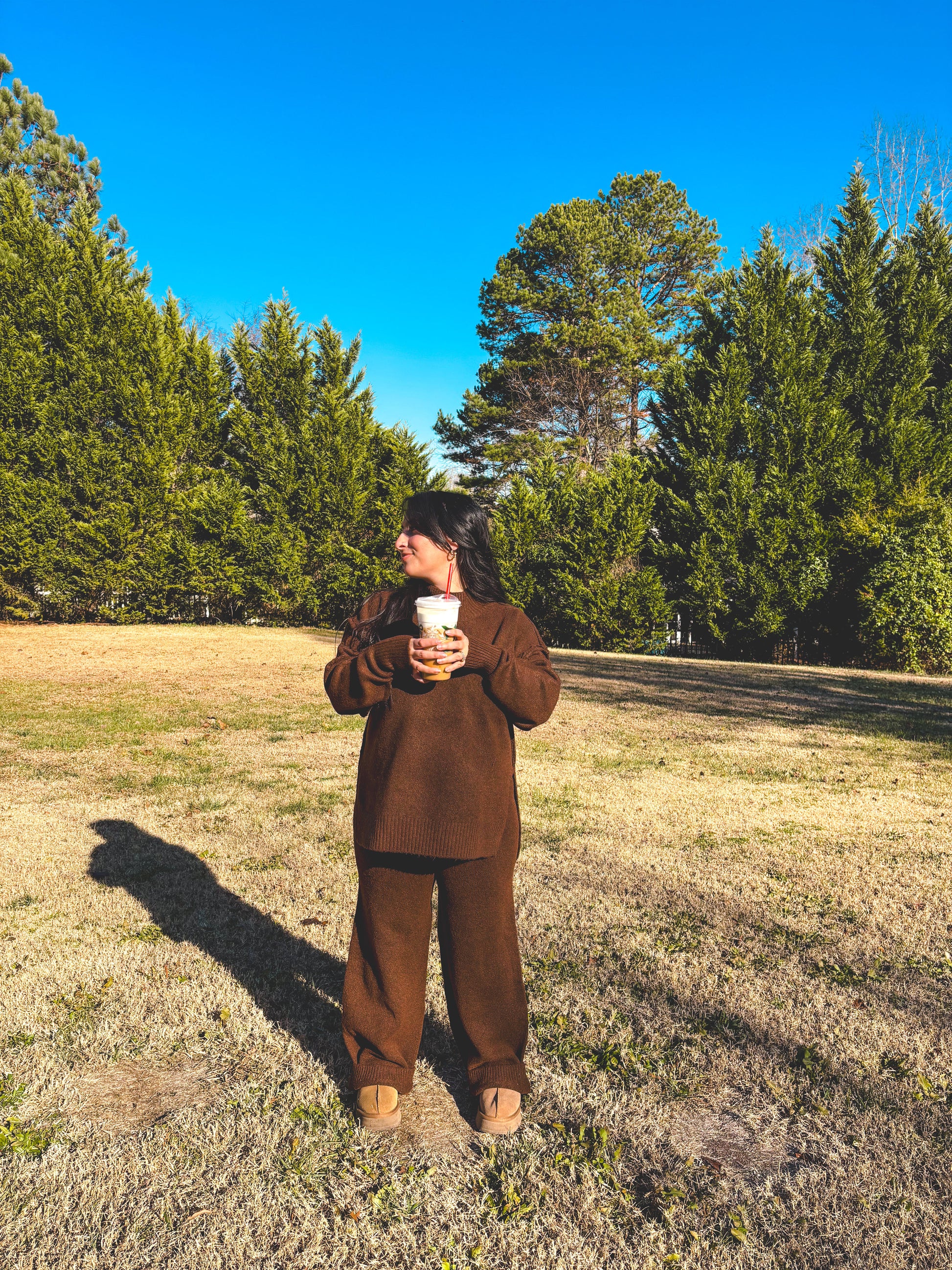 Person in a brown outfit pant set standing in a field with trees under a clear blue sky