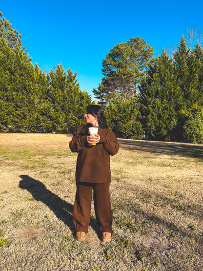 Person in a brown outfit pant set standing in a field with trees under a clear blue sky