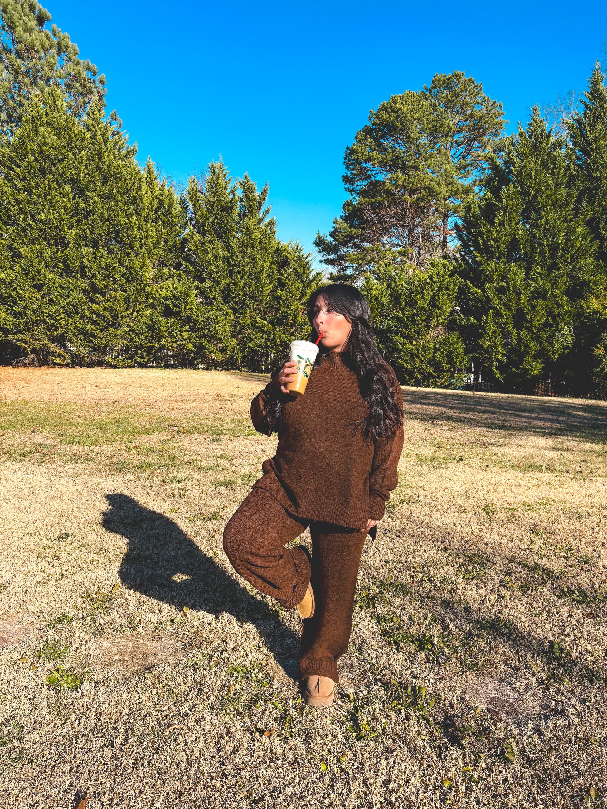 Person in brown outfit set holding a coffee cup in a park with trees and clear sky